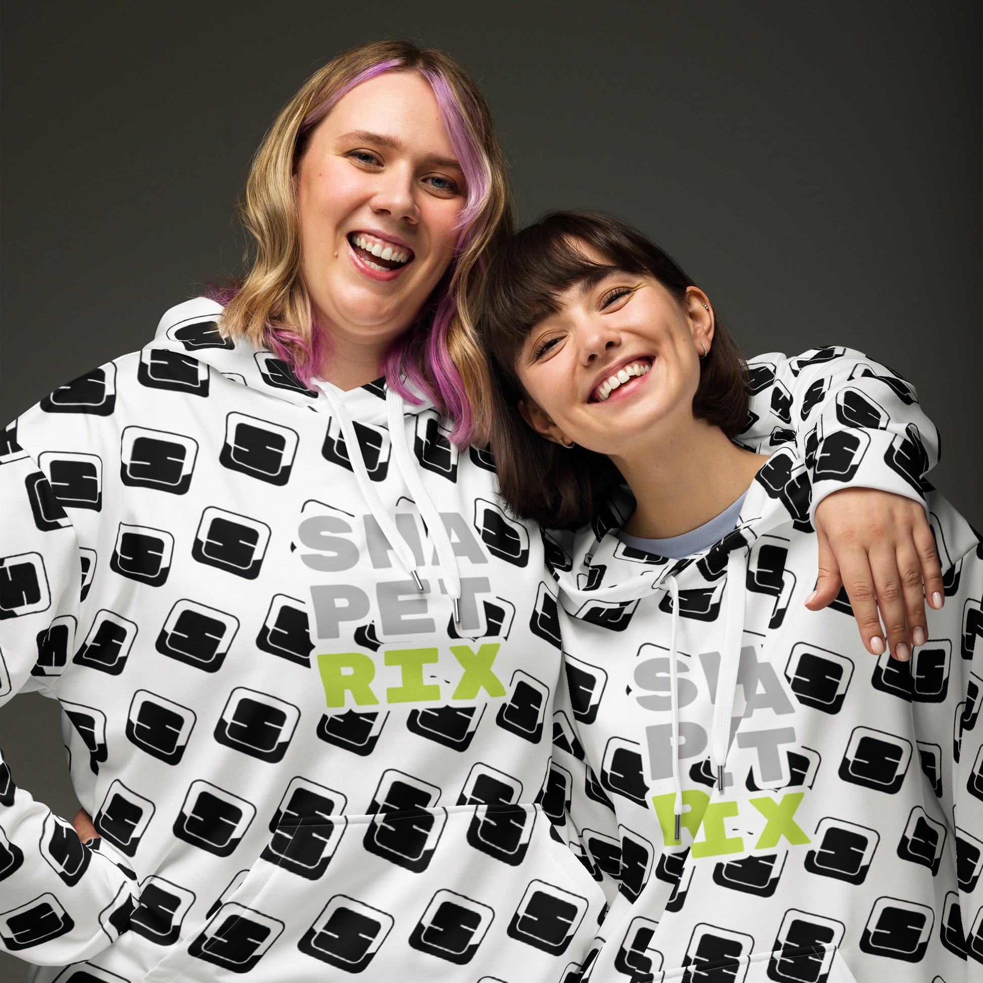 This image captures two joyful women wearing matching white hoodies adorned with the black Shapetrix 'S' logo pattern and bold lime green text spelling "SHAPETRIX" across the chest. The woman on the left has shoulder-length hair with pink highlights and is smiling broadly. The woman on the right, with a short brown bob and bangs, also displays a big, cheerful smile. They are embracing each other in a friendly pose, clearly enjoying the moment together against a grey studio background.