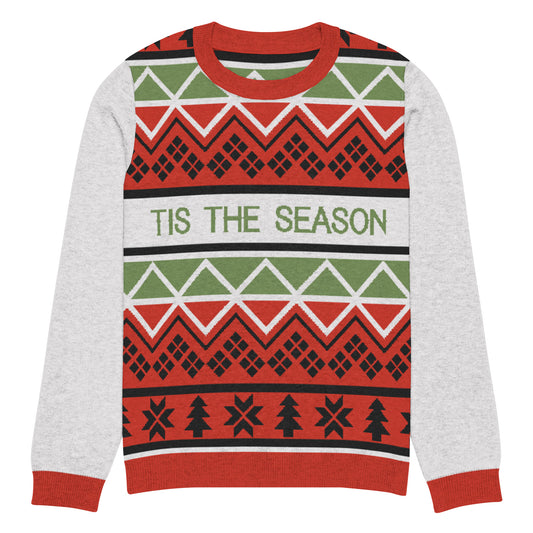 A festive holiday sweater with a crew neckline and contrasting gray raglan sleeves. The design features traditional Christmas patterns in red, green, and white. Horizontal bands include geometric diamonds, zigzags, and rows of Christmas trees, with 'TIS THE SEASON' written in bold green letters across the chest. 