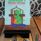 A vibrant photo displaying the cover of "Darren's Christmas Adventure," a storytime coloring book. The cover features Darren, a cheerful green dinosaur, standing in front of a fireplace, happily placing a piece of the Nativity scene on the mantle. Beside the book, on a wooden stand, is an array of scattered crayons, adding a playful and creative touch to the scene set against a patterned backdrop and a lush plant.