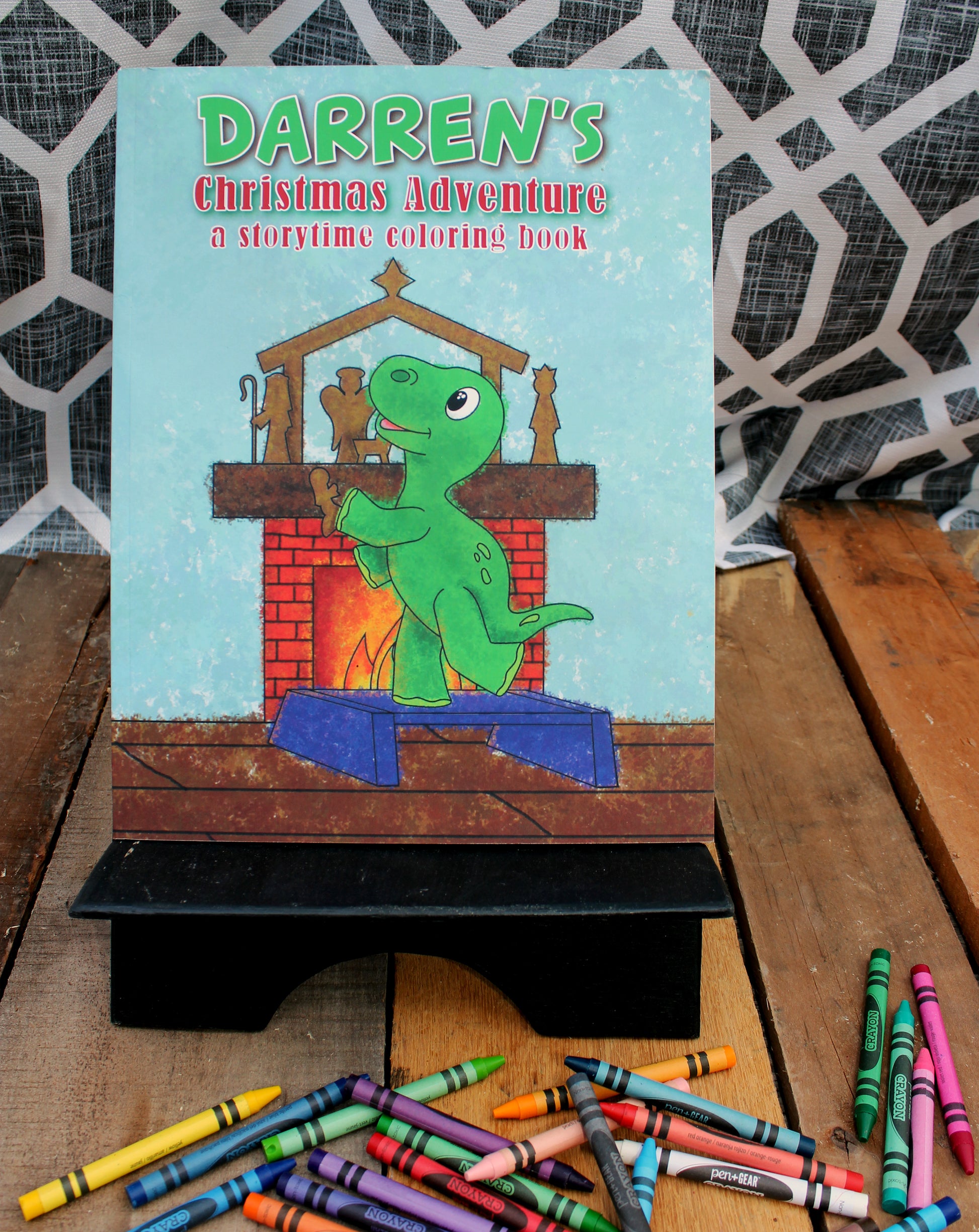 A vibrant photo displaying the cover of "Darren's Christmas Adventure," a storytime coloring book. The cover features Darren, a cheerful green dinosaur, standing in front of a fireplace, happily placing a piece of the Nativity scene on the mantle. Beside the book, on a wooden stand, is an array of scattered crayons, adding a playful and creative touch to the scene set against a patterned backdrop and a lush plant.