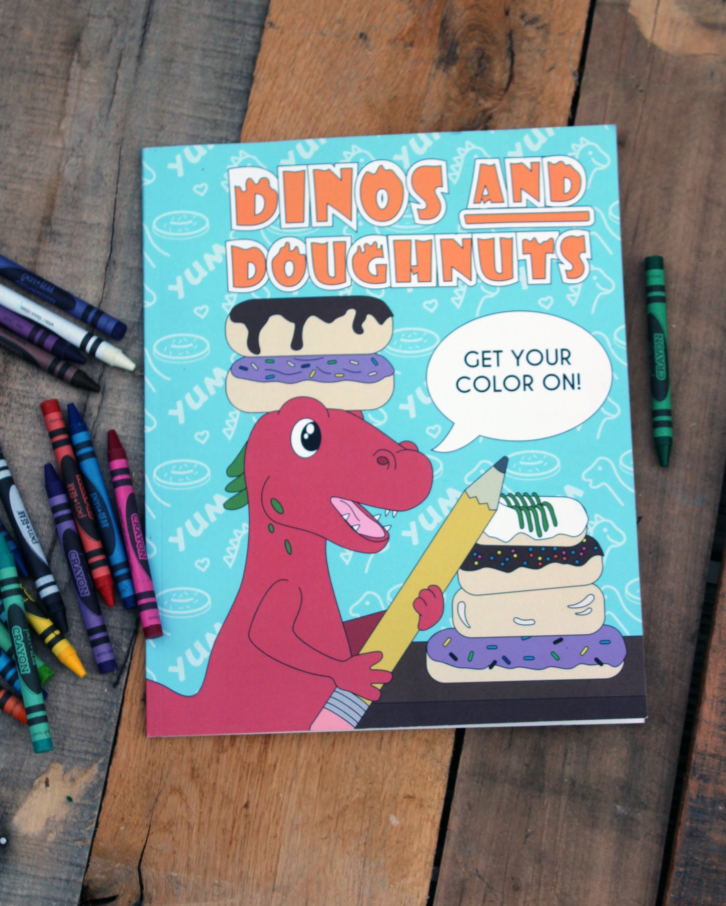 A vibrant photo showcasing the "Dinos and Doughnuts" coloring book resting on a rustic wooden surface. The cover features a cheerful red dinosaur holding a giant pencil next to a stack of colorful, frosted doughnuts. A speech bubble reads "GET YOUR COLOR ON!" Crayons are scattered around the book, emphasizing the creative and fun theme.