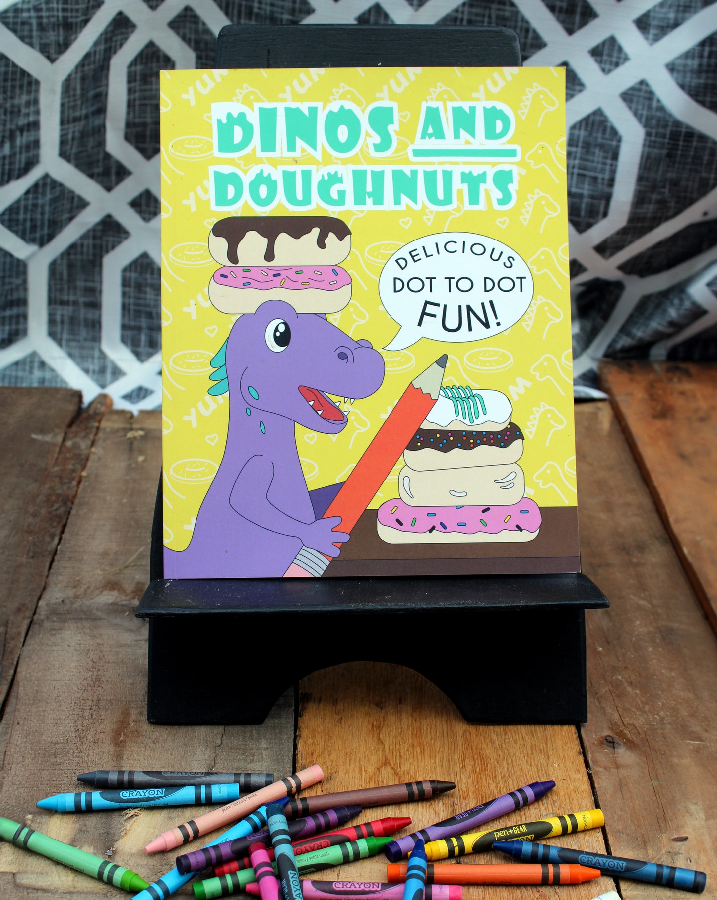 A colorful photo showcasing the "Dinos and Doughnuts" dot-to-dot activity book displayed on a black stand. The cover features a cheerful purple dinosaur holding a giant pencil next to a stack of doughnuts with vibrant icing and sprinkles. The text reads "Delicious Dot to Dot Fun!" against a bright yellow background patterned with doughnuts and the word "YUM." Crayons in various colors are scattered on the wooden surface below, adding a creative and playful vibe to the scene.