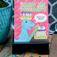 A vibrant photograph of the "Dinos and Doughnuts" activity book displayed on a small black stand. The book cover features a cheerful blue dinosaur holding a green pencil, surrounded by colorful doughnuts and the playful text "Trace the Treats!" in a speech bubble. The background is a lively pink pattern with "YUM" and doughnut illustrations. Crayons are scattered on the wooden surface below, and a bright blue potted plant adds a decorative touch to the setup, making the scene inviting and fun.