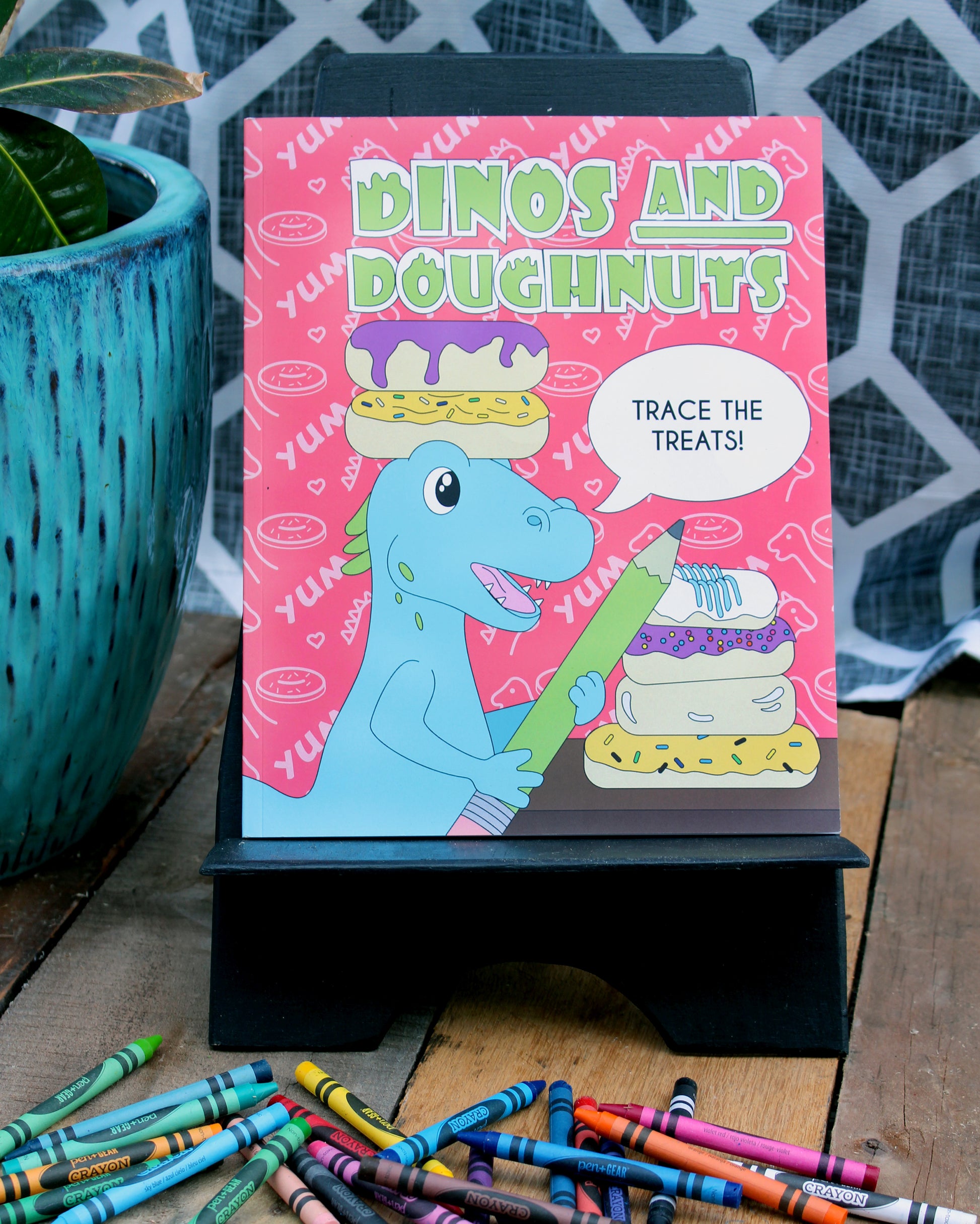 A vibrant photograph of the "Dinos and Doughnuts" activity book displayed on a small black stand. The book cover features a cheerful blue dinosaur holding a green pencil, surrounded by colorful doughnuts and the playful text "Trace the Treats!" in a speech bubble. The background is a lively pink pattern with "YUM" and doughnut illustrations. Crayons are scattered on the wooden surface below, and a bright blue potted plant adds a decorative touch to the setup, making the scene inviting and fun.