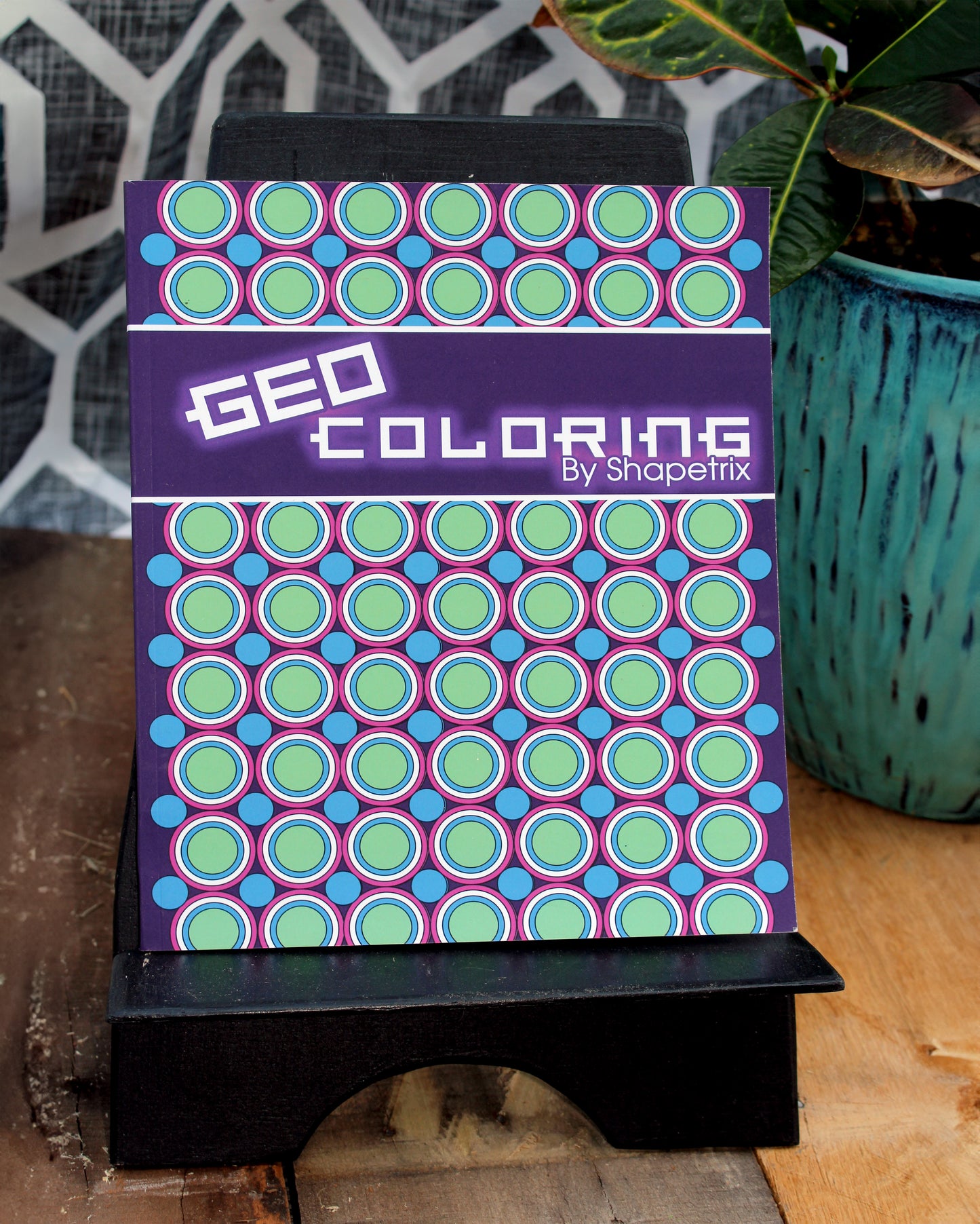 Geo Coloring Book by Shapetrix displayed on a wooden stand, featuring a vibrant geometric pattern with green, blue, and purple circular designs on the cover. A modern and creative coloring book perfect for adults seeking relaxation and mindfulness.