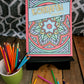 A photo of the "Kolidafun Volume 1" coloring book displayed upright on a black stand. The cover features a colorful mandala design with a central blue flower surrounded by pastel shades of green, pink, and purple. A light blue banner with yellow text spans diagonally across the cover. The setup includes vibrant colored pencils scattered on a rustic wooden surface, with some pencils placed in a green cup on the left. The background features a geometric black-and-white patterned fabric, adding a stylish touch