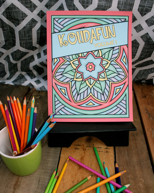 A photo of the "Kolidafun Volume 1" coloring book displayed upright on a black stand. The cover features a colorful mandala design with a central blue flower surrounded by pastel shades of green, pink, and purple. A light blue banner with yellow text spans diagonally across the cover. The setup includes vibrant colored pencils scattered on a rustic wooden surface, with some pencils placed in a green cup on the left. The background features a geometric black-and-white patterned fabric, adding a stylish touch