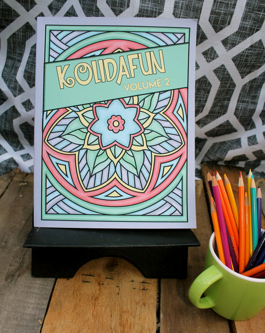 A photo of the "Kolidafun Volume 2" coloring book displayed on a wooden surface with a black stand. The cover features a colorful mandala design with a blue flower at the center, surrounded by geometric patterns. The title, "Kolidafun Volume 2," is written on a diagonal  banner across the top. To the right of the book, a green mug holds an assortment of colorful pencils, enhancing the creative theme of the image. A patterned black-and-white background adds a modern touch.