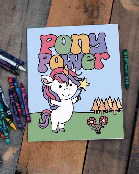 A vibrant cover for a coloring book displayed on a rustic wooden surface surrounded by colorful crayons. The illustration features a cheerful white unicorn with a rainbow mane and tail, holding a yellow star wand. Above the unicorn, the title "Pony Power" is written in bold, multicolored, playful letters. The background showcases a grassy landscape with golden trees, pink flowers, and a small mushroom under a clear blue sky. 