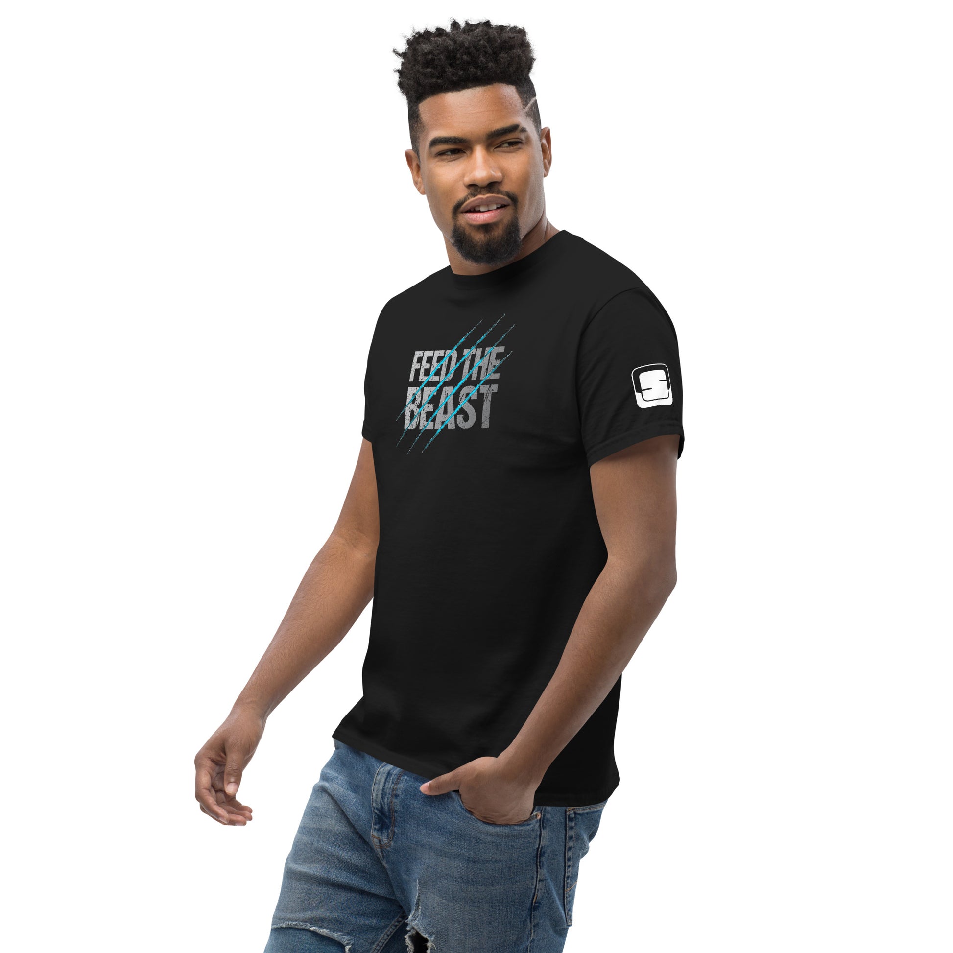 A confident young man with a short beard and stylish black hair, wearing a black t-shirt featuring the text 'FEED THE BEAST' in distressed white and blue with diagonal scratch graphics. He is paired with distressed blue jeans and poses with one hand on his hip, looking slightly to the side.