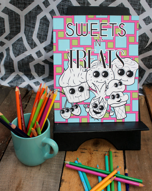 A playful coloring book titled "Sweets N' Treats" displayed on a wooden stand. The cover features cheerful dessert characters, including a cupcake, pineapple, ice cream cone, and cherries, all with large expressive eyes. The background showcases a vibrant checkered pattern in pink, blue, and green. In the foreground, colored pencils are scattered on the wooden table, with a teal mug holding additional pencils, creating a creative and inviting scene.