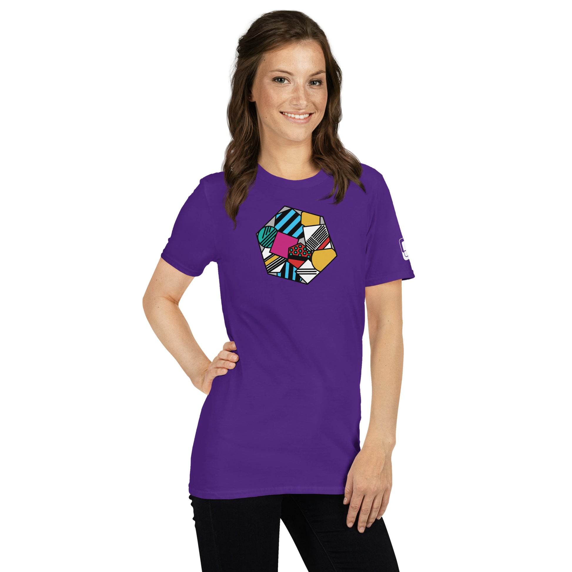 A cheerful Caucasian woman poses in a vibrant purple tee shirt adorned with a colorful geometric pattern in a hexagon shape. The design includes stripes, polka dots, and various textures in lively colors of blue, green, yellow, and red.