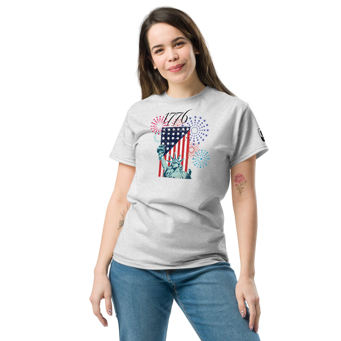 A young woman wearing a light gray t-shirt that features a vibrant Fourth of July graphic. The design prominently displays the Statue of Liberty with an American flag background, surrounded by colorful fireworks in shades of red, blue, and purple. Above the image, the year '1776' is highlighted, symbolizing American independence.