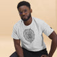 Focused man with glasses and a short beard wearing a heather grey t-shirt adorned with a central geometric lion head illustration, sporting a logo tag on the left sleeve, seated against a soft beige backdrop.