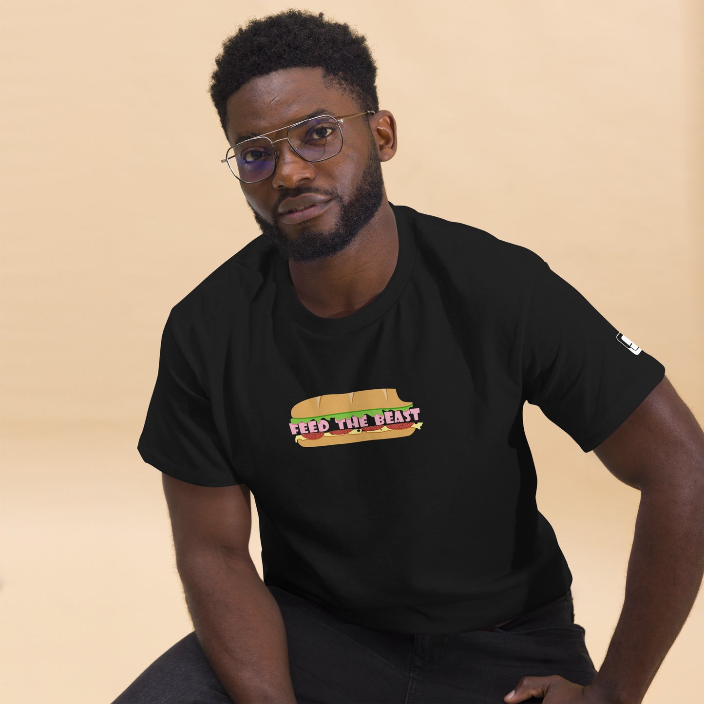 Confident young man wearing glasses and a black t-shirt that features a colorful design featuring the text 'FEED THE BEAST' as the deli meat inside the bun. He is seated, leaning forward slightly with a serious expression, looking directly at the camera. The background is a soft, warm beige tone, complementing his dark attire and focused demeanor.