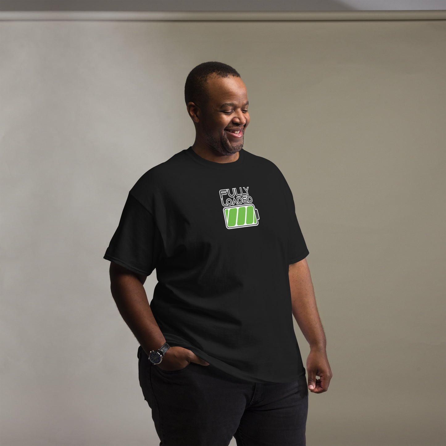 A cheerful African American man models a black t-shirt emblazoned with a green battery icon and the words "FULLY LOADED" in white. He stands casually with his right hand in his pocket and a warm smile, wearing black pants and a watch. The simple yet striking design of the t-shirt contrasts effectively against the soft gray background, highlighting the vibrant green graphic.