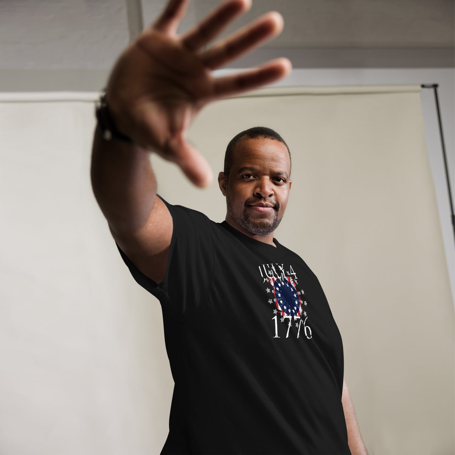 A middle-aged African American man poses confidently with his hand raised towards the camera, giving a stop gesture. He wears a blackt-shirt with a patriotic "July 4, 1776" design featuring a flag motif in red, white, and blue. His expression is serious yet friendly. The setting is a softly lit indoor environment, enhancing the focus on his expressive pose and the detailed graphic on his shirt.