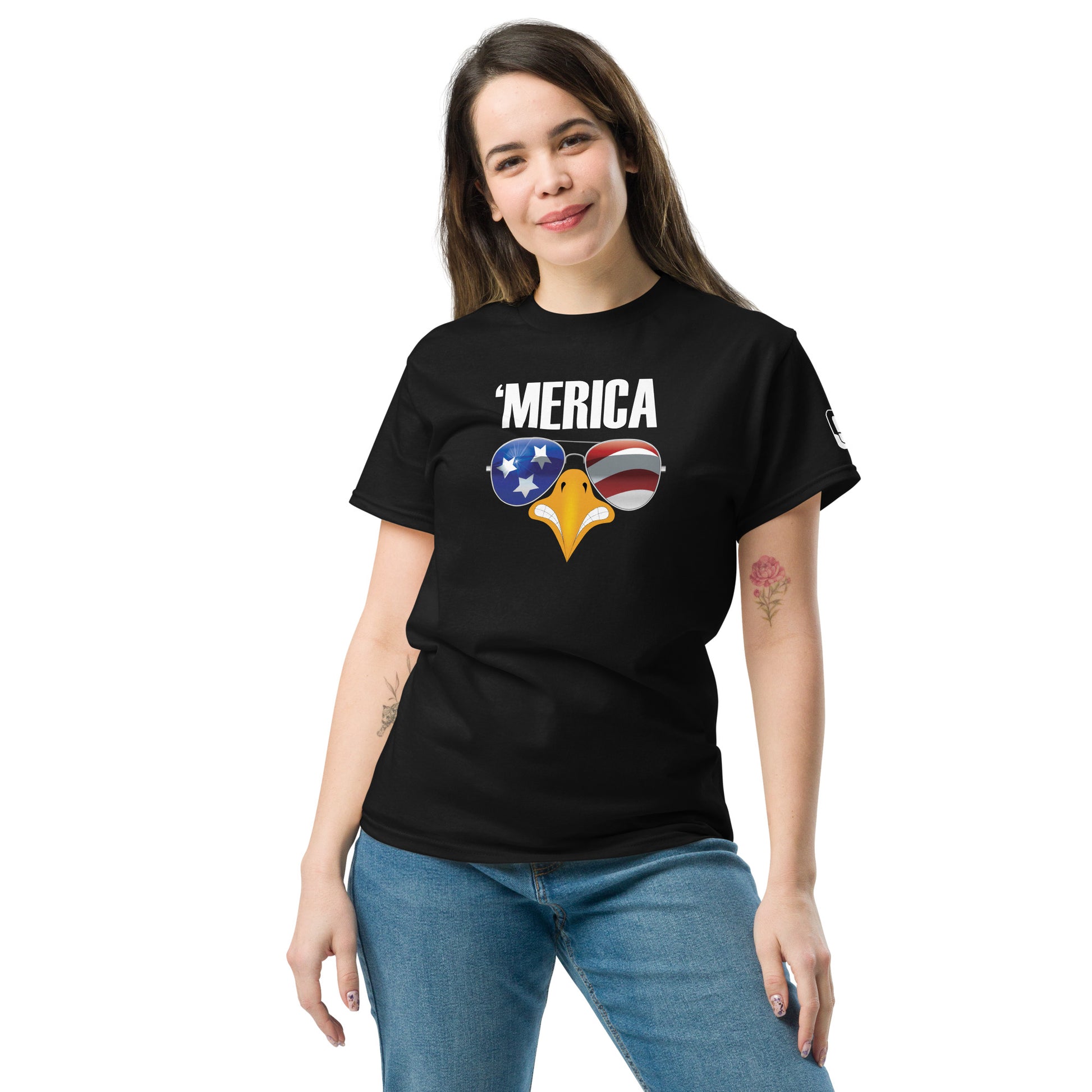 A young woman with a pleasant expression stands in a relaxed pose, wearing a black t-shirt featuring a strong graphic. The design includes the word "'MERICA" in white, above a stylized eagle's beak adorned with sunglasses reflecting the American flag—stars against a blue background on one lens and red and white stripes on the other. The fun and patriotic theme of the shirt is highlighted against the simple black backdrop of the t-shirt, making it a casual yet striking piece of apparel.