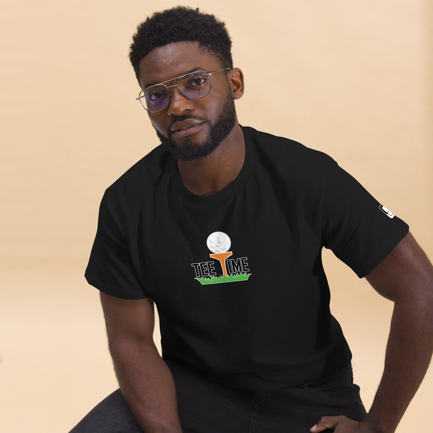 Man with glasses wearing a black short-sleeved t-shirt with a 'Tee Time' graphic showing a golf ball character on grass on the chest and a small logo patch on the sleeve, sitting against a beige background.