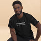 A focused young Black man with a neatly styled beard and short hair, wearing glasses, is depicted in a black t-shirt that reads "On Tuesdays We Wear T-Shirts" in simple white font. His pose is slightly leaned forward, and he looks directly at the viewer with a serious and engaging expression. The warm-toned background enhances the casual and modern feel of the image. His t-shirt is accessorized with a small logo on the sleeve, adding a subtle detail to his sleek attire.