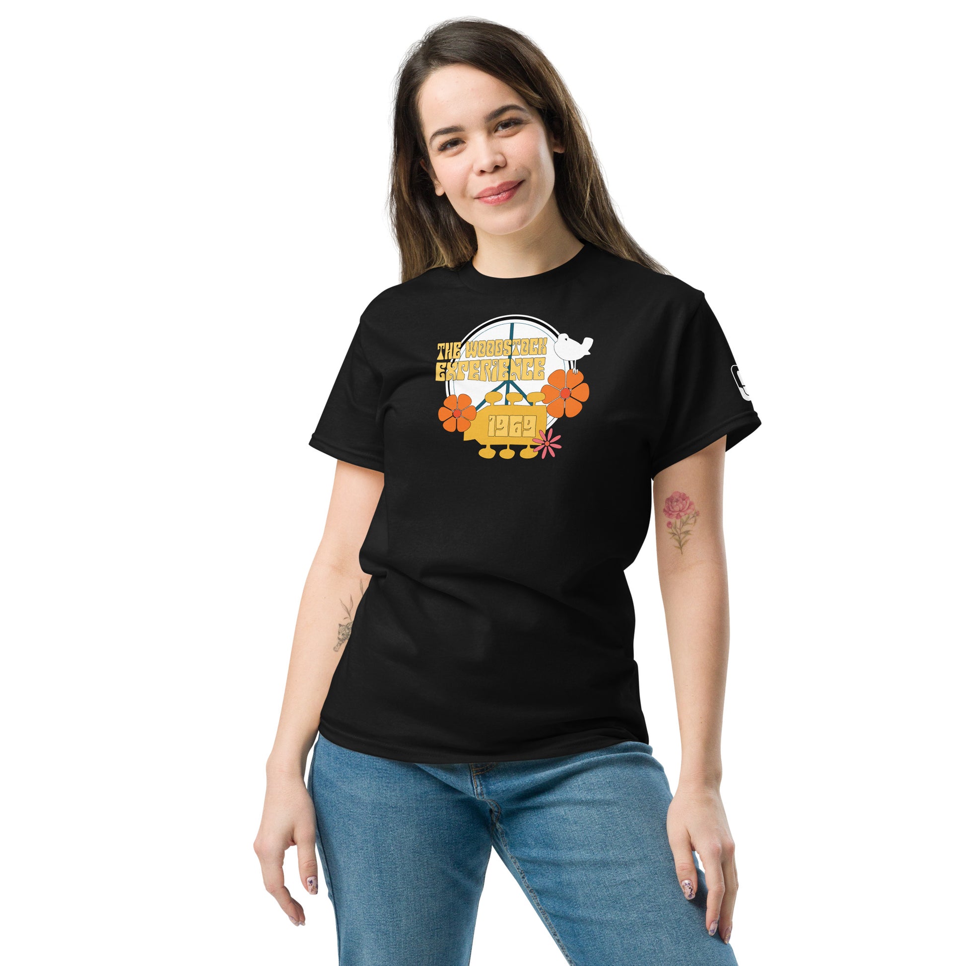 A young woman smiles gently while wearing a black t-shirt featuring a colorful and retro graphic design titled "The Woodstock Experience 1969". The design includes a peace symbol encircled by the event's name in vintage-style lettering, flanked by orange and pink flowers. A simple white dove perches on the lettering, adding a touch of peace symbolism. The shirt pairs casually with her blue jeans, creating a relaxed and fashionable ensemble that nods to the iconic music festival's spirit of peace and music.