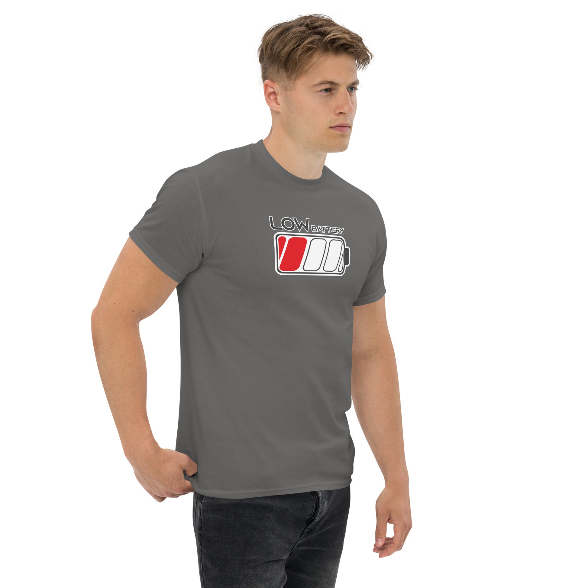 A young Caucasian man models a gray t-shirt printed with the design "LOW BATTERY" in stylized grey lettering with a white outline. The design features a partially filled battery icon with the leftmost cell highlighted in red. The man stands in a relaxed pose with his hands casually placed on his hip, gazing slightly off to the side against a neutral background.