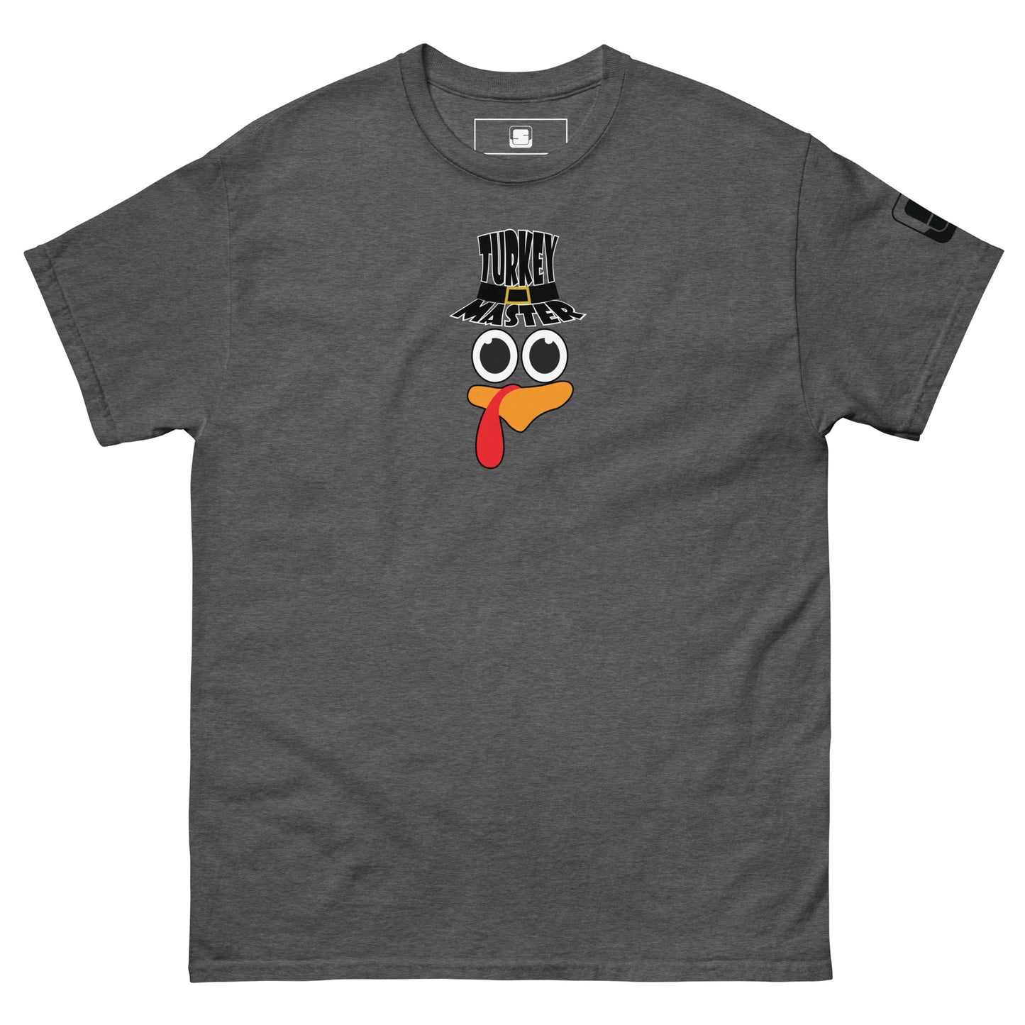 A dark heather t-shirt featuring a playful graphic design of a turkey face with eyes, an orange beak, and a red snood. Above the face, the text reads "TURKEY MASTER" in bold, stylized letters. The text is in the shape of a pilgrims hat. On the left sleeve, there's a rectangular logo patch, adding a unique touch. The shirt is laid out flat, showcasing the entire design clearly.