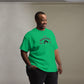 A cheerful African American man wears a vibrant green t-shirt featuring the playful phrase "I'm Your Huckleberry" in bold black lettering, arched over a stylized black cowboy hat motif. He stands in a relaxed pose, smiling warmly, set against a soft grey background that highlights the vivid color and fun design of the shirt.