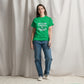 A young Caucasian woman stands confidently against a draped white curtain background, wearing a bright green t-shirt with the phrase "DON'T GET YOUR TINSEL IN A TANGLE" in bold teal text. She pairs her vibrant shirt with classic blue jeans and white sneakers, presenting a casual and relaxed look. 