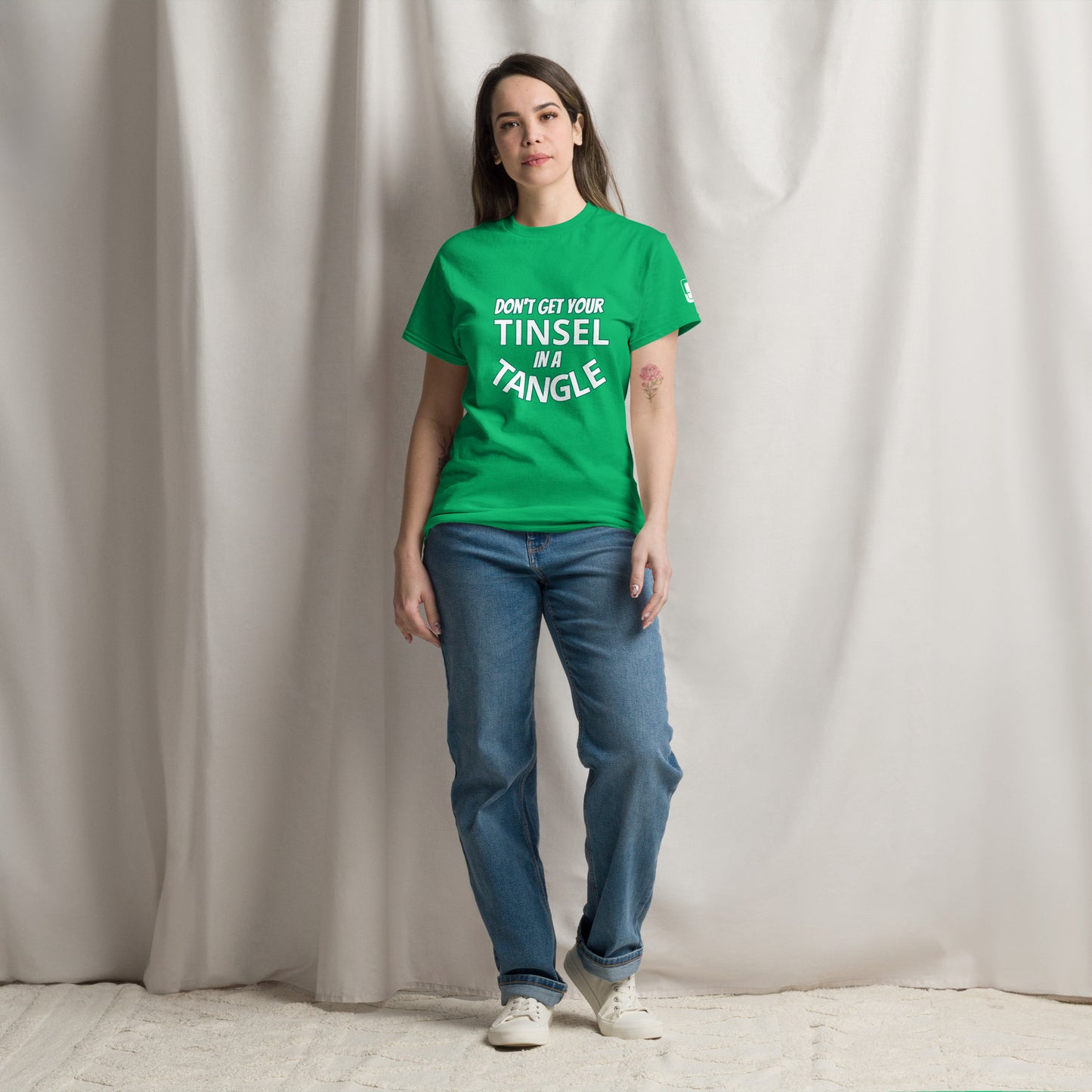 A young Caucasian woman stands confidently against a draped white curtain background, wearing a bright green t-shirt with the phrase "DON'T GET YOUR TINSEL IN A TANGLE" in bold teal text. She pairs her vibrant shirt with classic blue jeans and white sneakers, presenting a casual and relaxed look. 