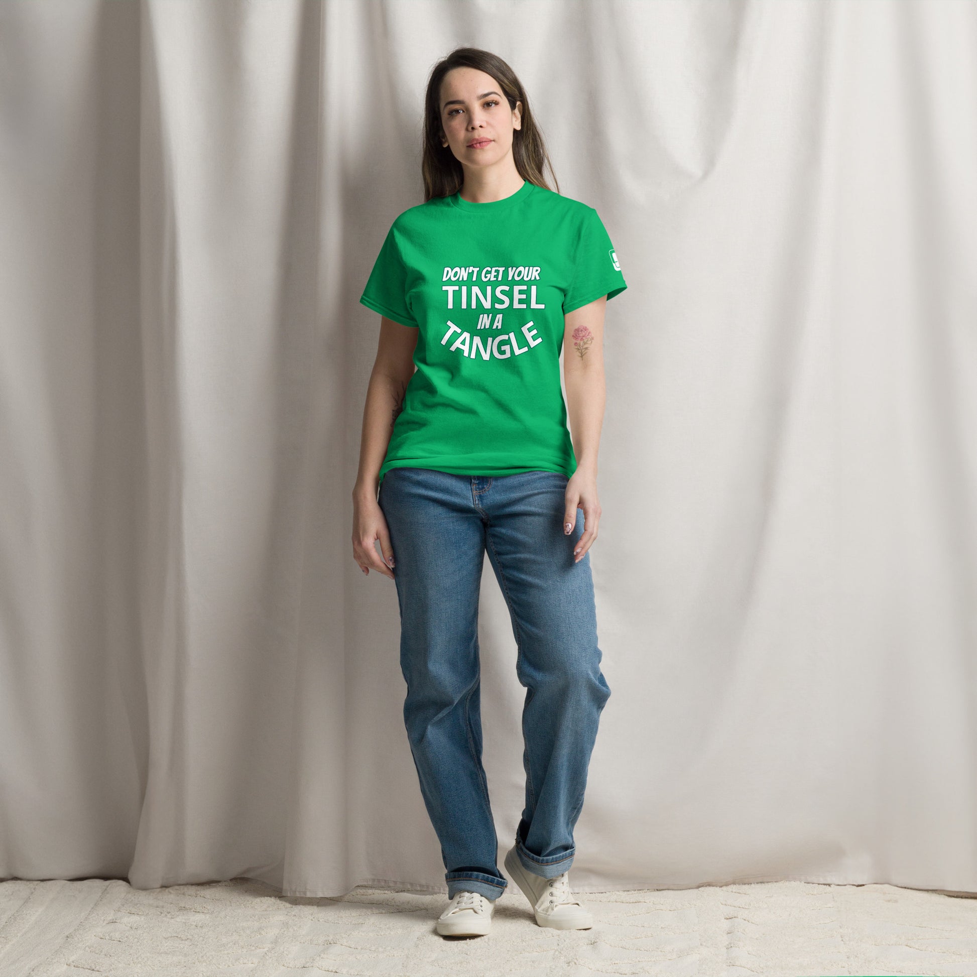 A young Caucasian woman stands confidently against a draped white curtain background, wearing a bright green t-shirt with the phrase "DON'T GET YOUR TINSEL IN A TANGLE" in bold teal text. She pairs her vibrant shirt with classic blue jeans and white sneakers, presenting a casual and relaxed look. 