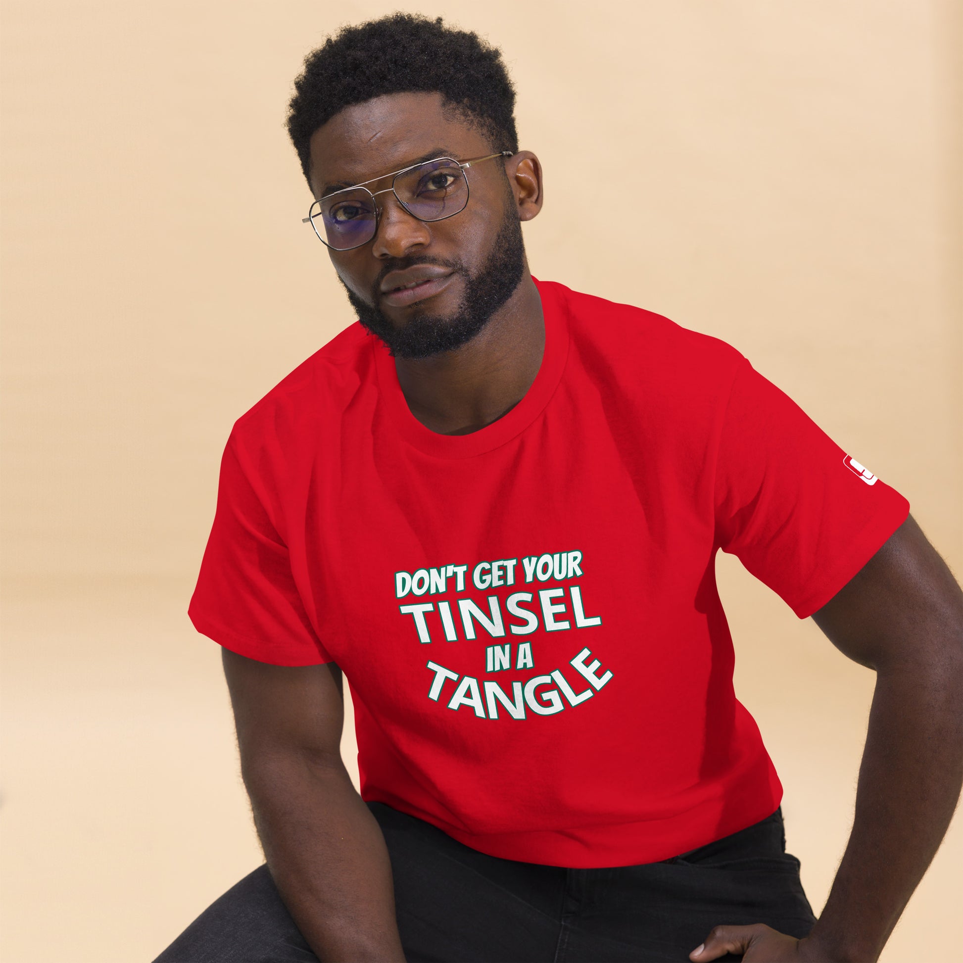 A young African American man confidently poses in a vivid red t-shirt that reads "DON'T GET YOUR TINSEL IN A TANGLE" in bold white letters. He wears stylish eyeglasses and sports a well-groomed beard, complementing his serious yet approachable expression. The soft beige background highlights his attire and adds warmth to the overall scene.