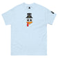 A light-blue t-shirt featuring a playful graphic design of a turkey face with eyes, an orange beak, and a red snood. Above the face, the text reads "TURKEY MASTER" in bold, stylized letters. The text is in the shape of a pilgrims hat. On the left sleeve, there's a rectangular logo patch, adding a unique touch. The shirt is laid out flat, showcasing the entire design clearly.