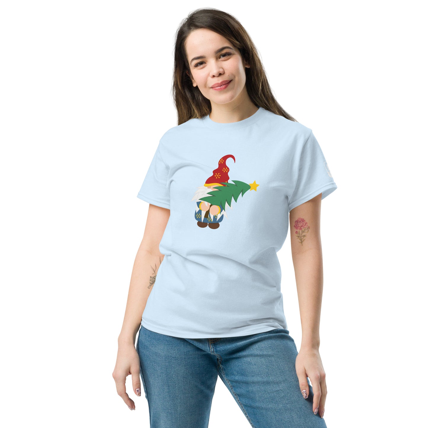 A woman in a light blue t-shirt that features a cheerful gnome holding a Christmas tree. The gnome sports a long white beard and a red hat adorned with golden snowflakes, holding a green Christmas tree with a star on top. She poses casually with her hand on her hip, smiling gently. The simple background of the t-shirt allows the colorful design to stand out.