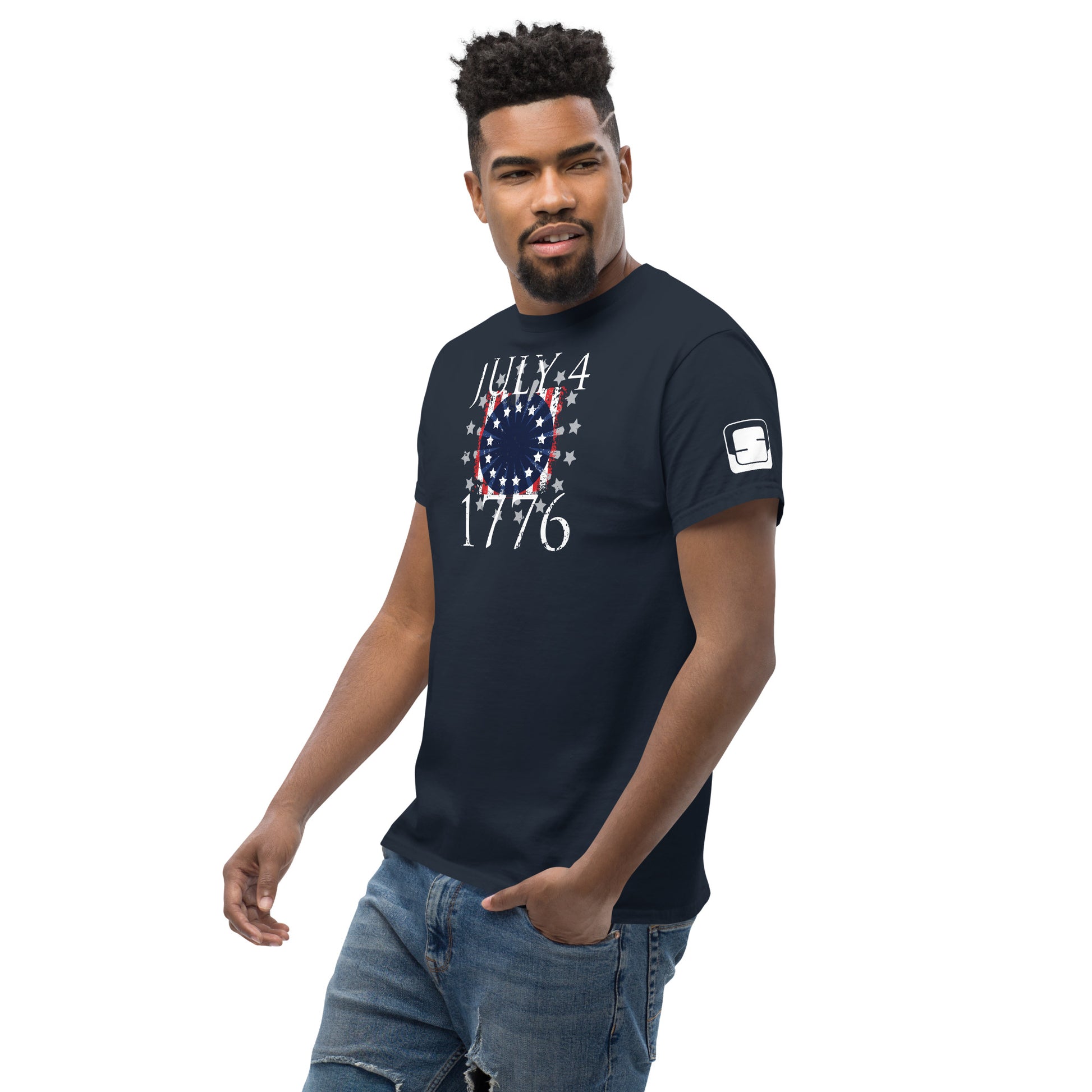 A stylish young African American man sports a dark navy t-shirt featuring a festive "July 4, 1776" design in red, white, and blue that mimics the appearance of a United States flag. He poses casually with one hand in his jean pocket, exuding confidence and a relaxed demeanor. His shirt, perfect for Independence Day celebrations, adds a patriotic flair to his modern, laid-back outfit.