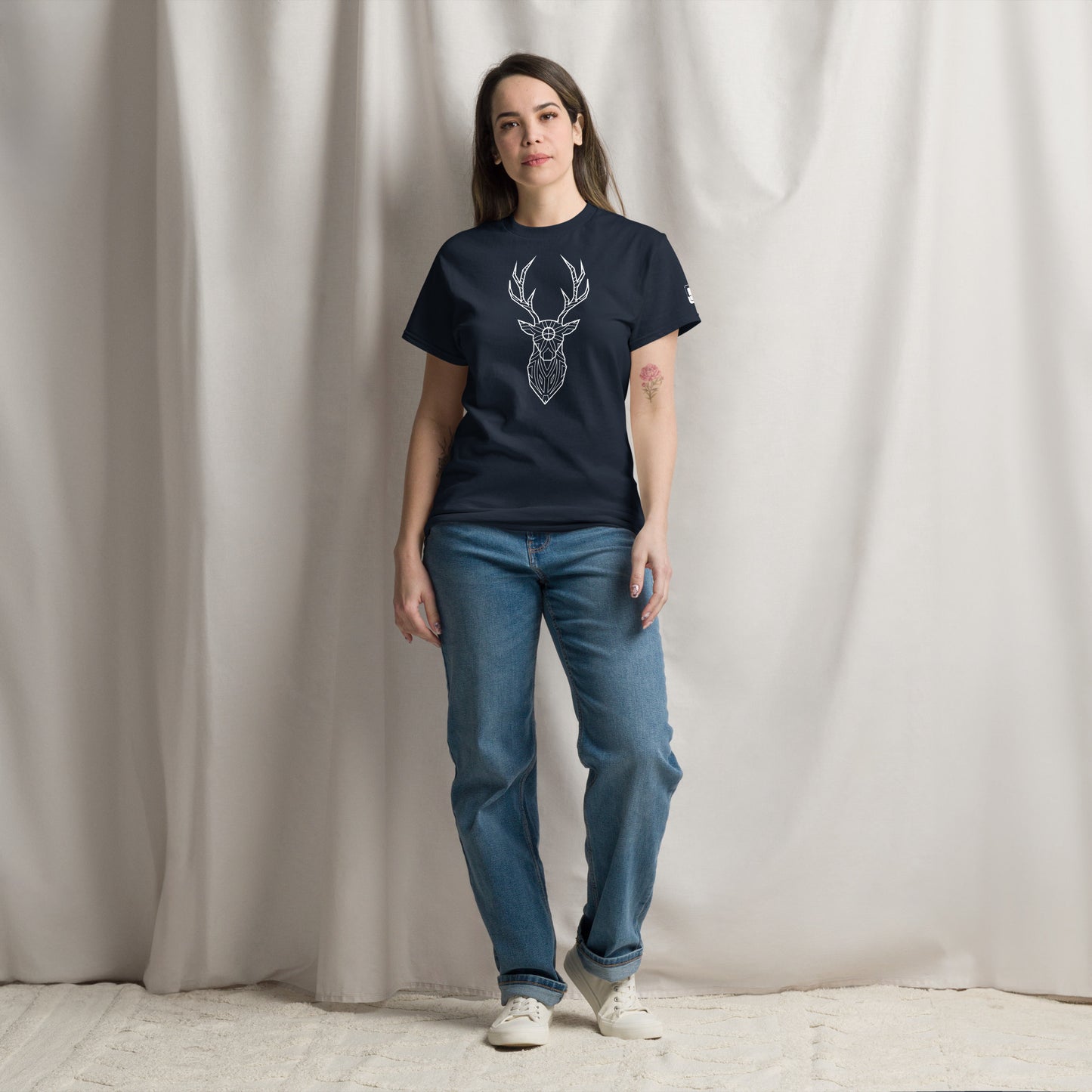 A young Caucasian woman stands relaxed in a studio, wearing a navy t-shirt with a white deer head design, paired with comfortable jeans.