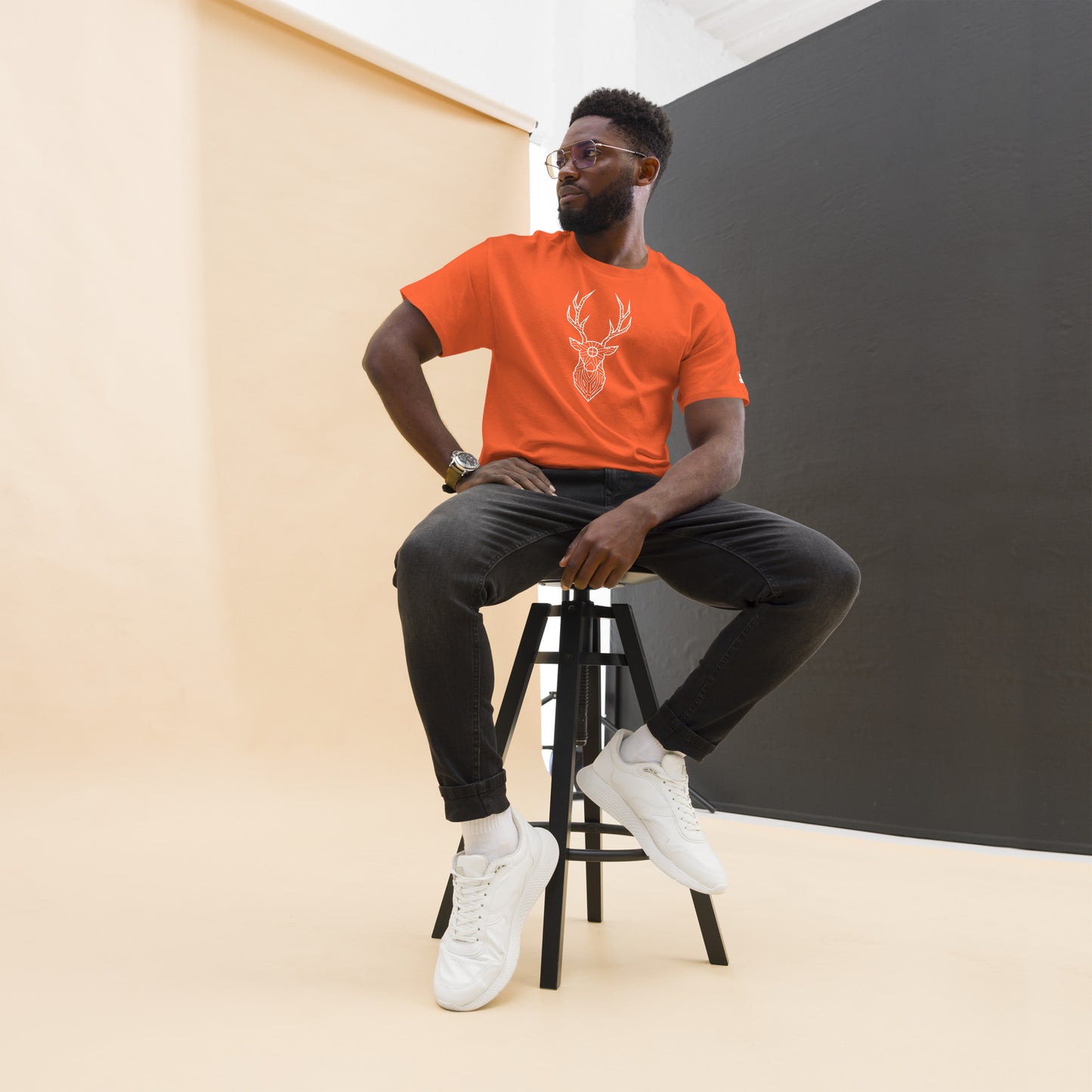 A confident African American man posing on a stool, wearing an orange t-shirt with a white geometric deer design, paired with dark jeans.