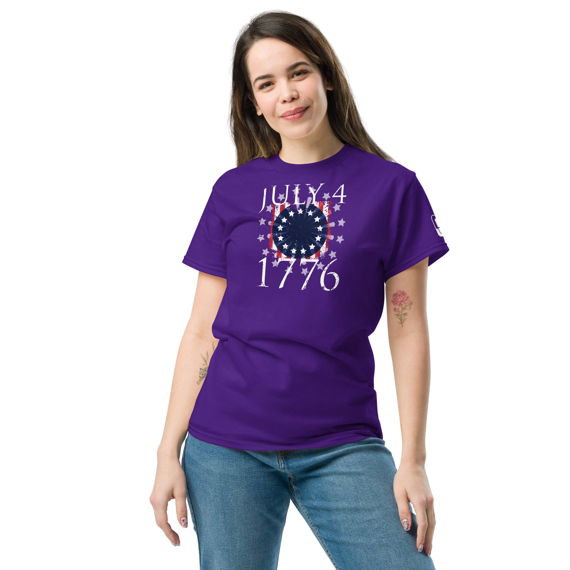 A young woman stands smiling, wearing a vibrant purple t-shirt that features a patriotic design celebrating Independence Day. The shirt displays a central graphic with "July 4, 1776" encircled by stars and stripes in white, symbolizing the American flag. The simple yet stylish design conveys a festive and historical theme. She pairs the tee with light blue jeans and appears relaxed and casual, perfect for a celebratory occasion.