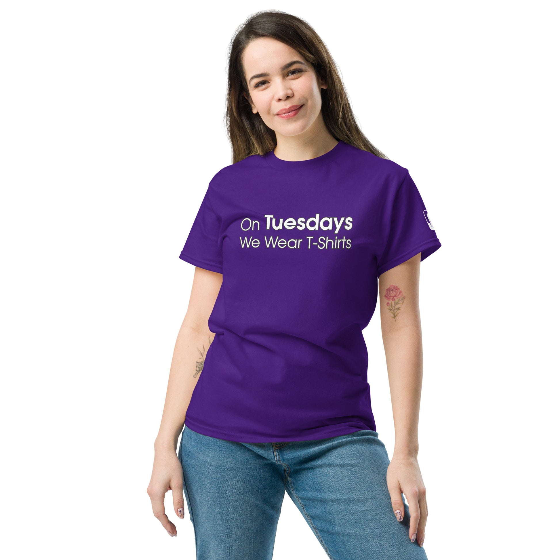 A young woman with medium-length brown hair, wearing a vibrant purple t-shirt that reads "On Tuesdays We Wear T-Shirts" in white text. She is smiling and standing casually with one hand on her hip, the other hanging loosely at her side. She is also wearing light blue jeans and sports a subtle tattoo on her right arm. The scene is set against a plain, light background, emphasizing the cheerful and relaxed demeanor of the subject.