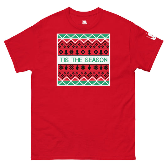 A bright red t-shirt featuring a large, festive design that resembles a Christmas sweater pattern. The design includes multiple rows of geometric and star patterns in green, white, and black, centered around the phrase 'TIS THE SEASON' in bold, green letters on a white background.