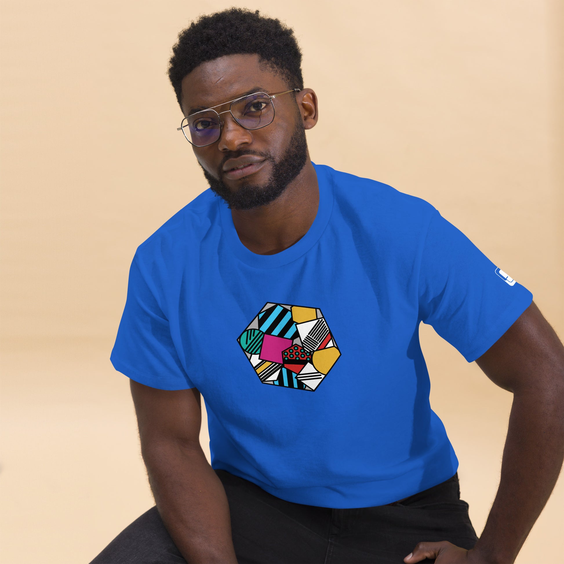 A young Black man in stylish glasses, posing confidently while seated. He wears a striking royal blue t-shirt featuring a colorful geometric design on the chest. The design is a hexagon filled with vibrant patterns including stripes, dots, and abstract shapes in shades of teal, pink, yellow, and white, set against a contrasting black background within the hexagon. His relaxed, yet stylish demeanor complements the modern and artistic flair of the t-shirt.