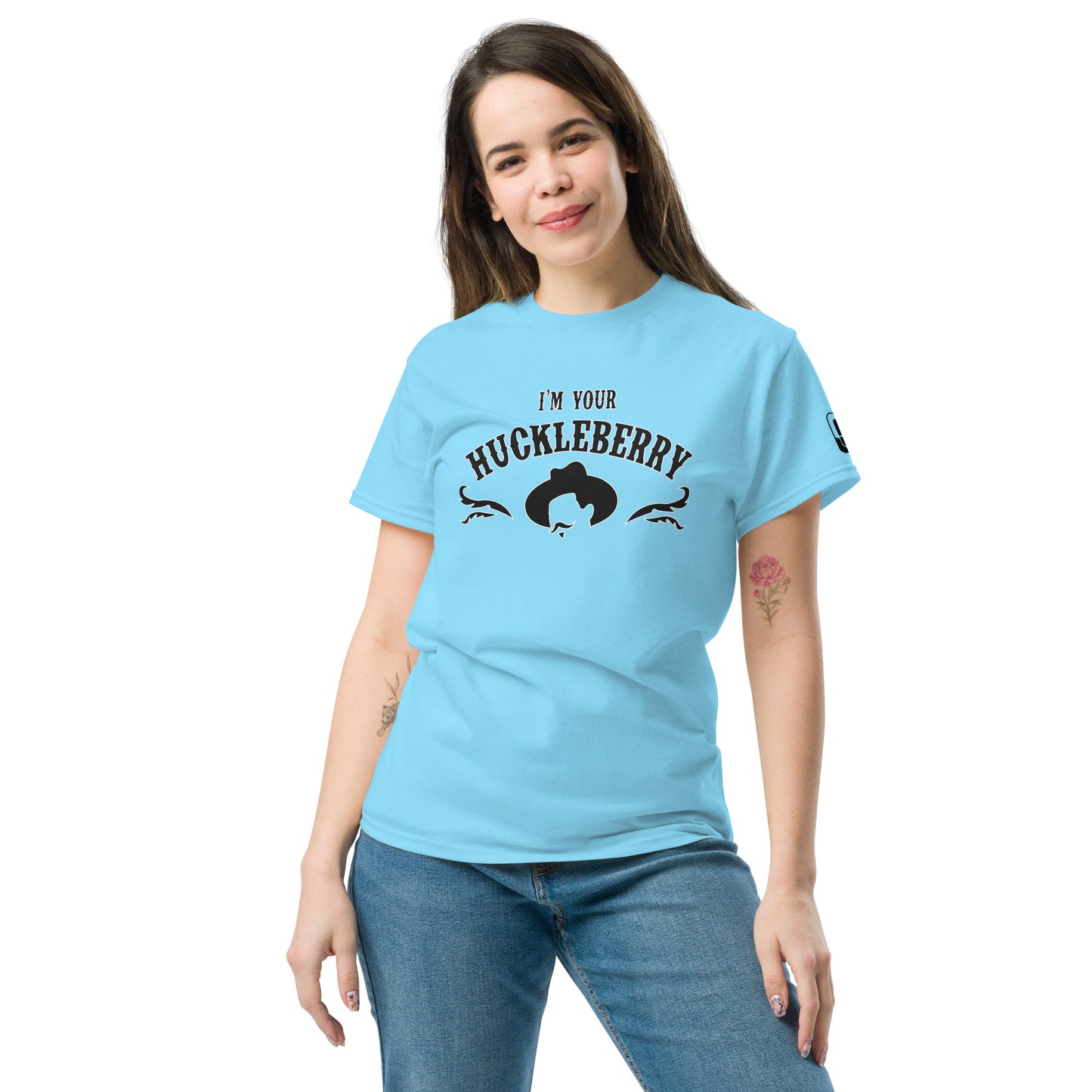 A young Caucasian woman models a light blue t-shirt that features the phrase 'I'm Your Huckleberry' in bold black letters arched over a stylized design. The design includes a black silhouette of a man's head with a cowboy hat, framed by intricate black filigree that adds some detail. She stands relaxed, with a slight smile, wearing the t-shirt paired with jeans.