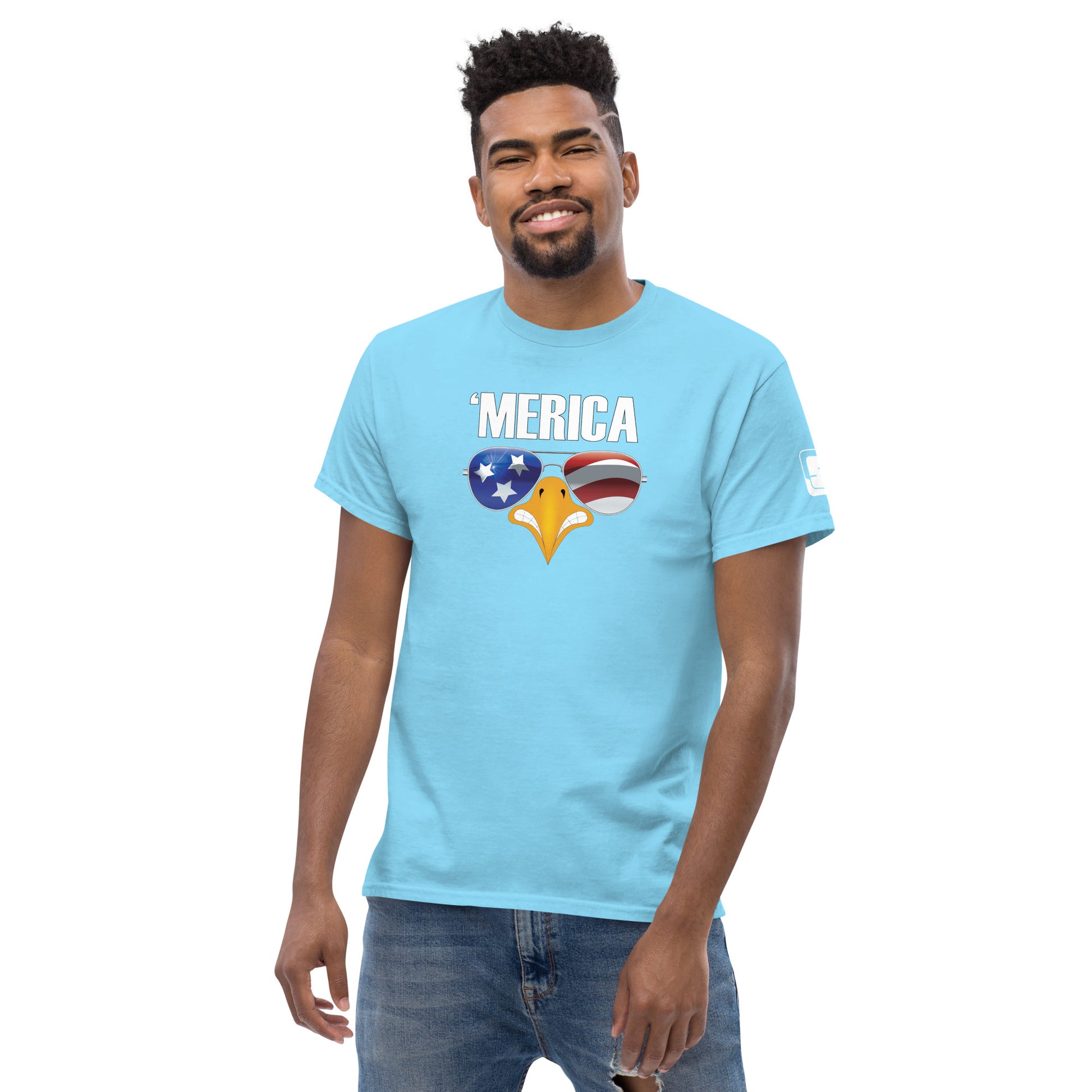 A cheerful young man with a friendly smile models a light blue t-shirt that features a bold patriotic design. The shirt displays the word "'MERICA" in white letters, with a graphic of an eagle's beak adorned with sunglasses reflecting a stylized American flag—stars on a blue background on the left lens and red and white stripes on the right. The design captures a fun and casual American spirit, perfect for patriotic events or casual wear.