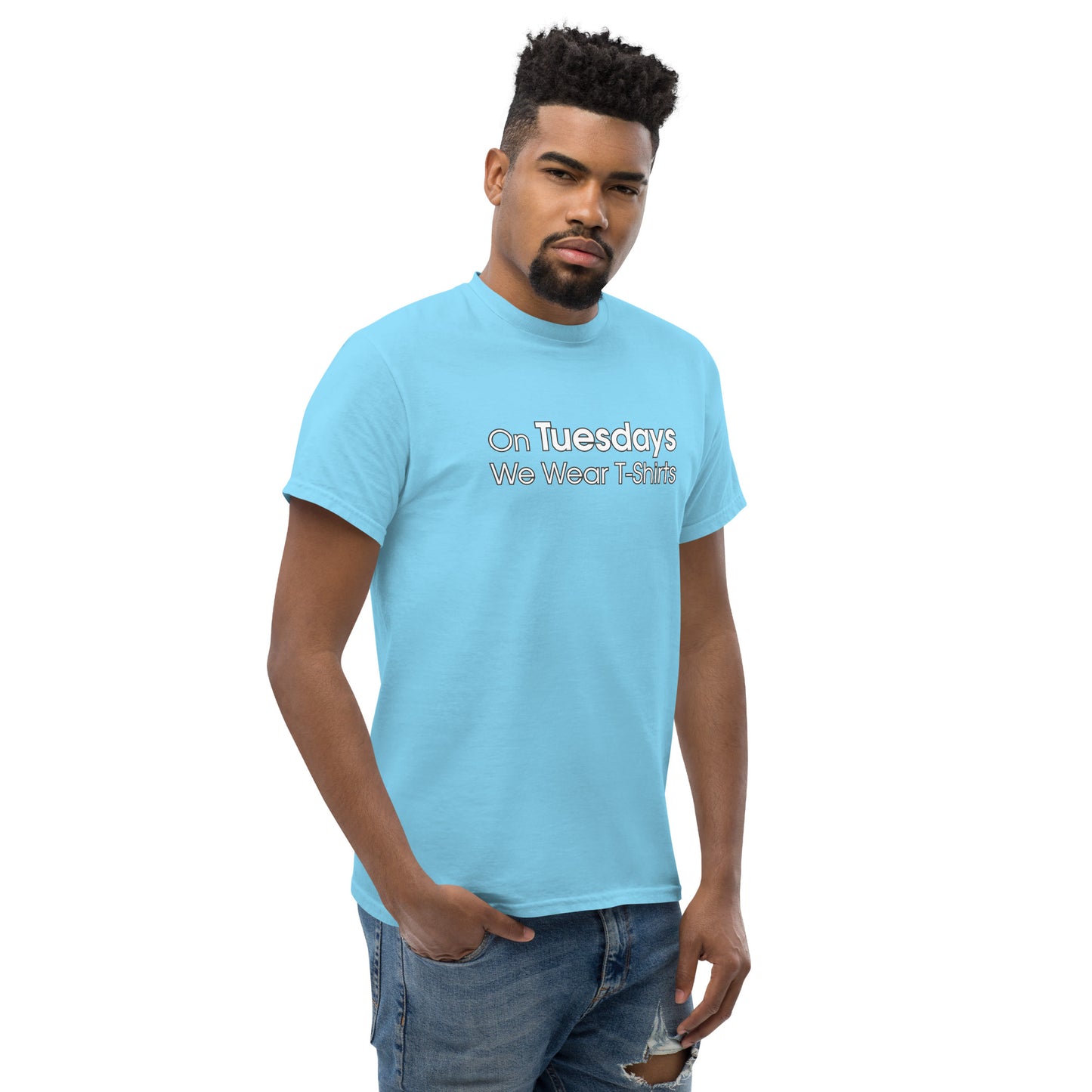 A confident young man with short, styled hair and a neatly groomed beard wears a light blue t-shirt featuring the phrase "On Tuesdays We Wear T-Shirts" in white lettering. He stands with one hand in his jeans pocket, gazing slightly to the side with a subtle, contemplative expression. His relaxed posture and casual ripped jeans complete a laid-back yet stylish look. The background is plain, focusing attention on him and the message on his shirt.