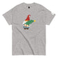 A light gray t-shirt featuring a playful gnome design. The gnome is adorned with a red hat embellished with golden snowflakes, a white beard, and is depicted carrying a green Christmas tree decorated with a yellow star on top. The t-shirt is casually styled with a small logo tag on the sleeve.