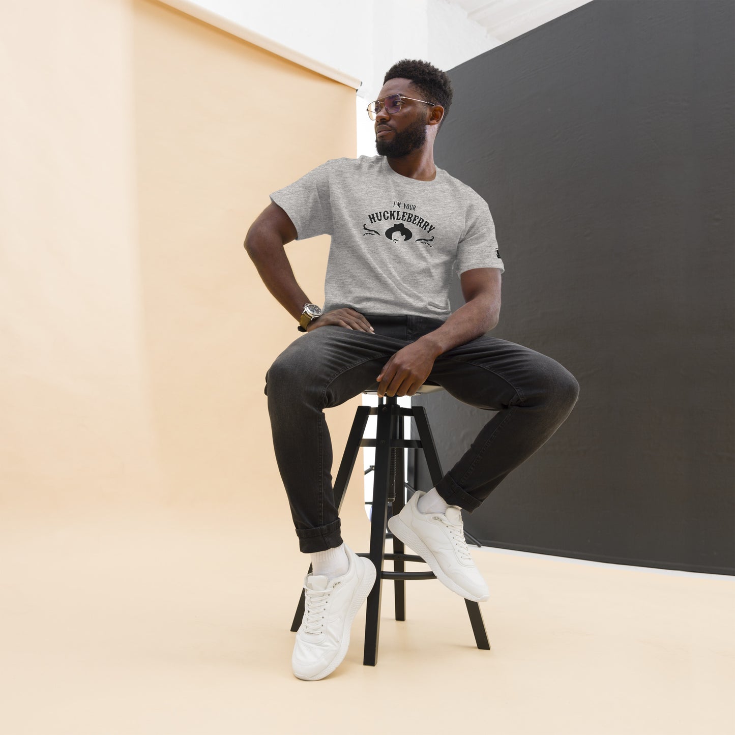 An African American man poses confidently, seated on a stool, modeling a heather grey t-shirt with the phrase 'I'm Your Huckleberry' printed in black. Under the text, a stylized black silhouette of a cowboy hat enhances the western motif. He completes his look with dark jeans, white sneakers, and stylish glasses, set against a contrasting backdrop of a light ochre wall and a dark grey panel.