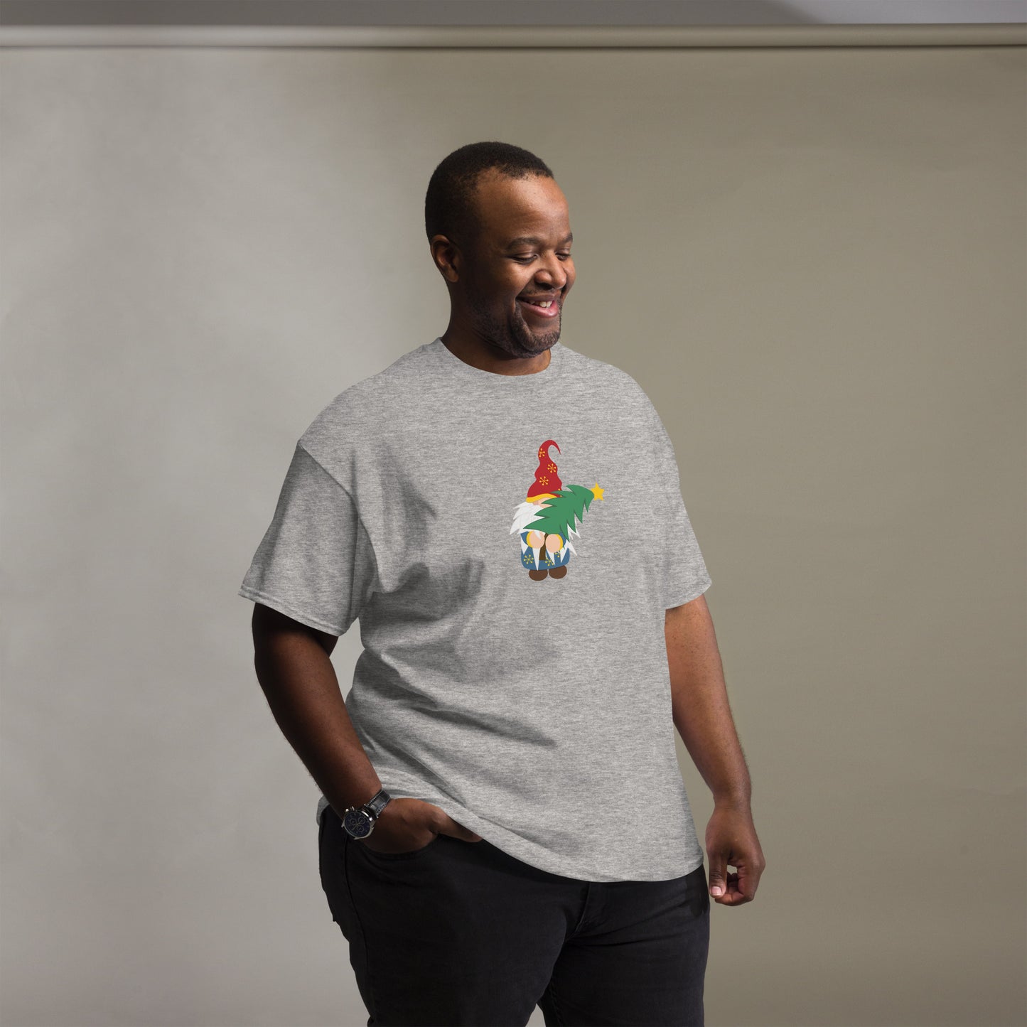 A man wearing a heather gray t-shirt featuring a festive gnome holding a Christmas tree. The gnome is adorned with a red hat decorated with gold snowflakes and has a long white beard. He is carrying a vibrant green Christmas tree topped with a yellow star. The man is smiling and appears relaxed, casually placing his hand in his pant pocket, set against a soft gray background.