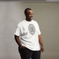 Smiling man wearing a white t-shirt with a detailed geometric lion head print on the front, paired with black pants, and a wristwatch, standing in a room with a grey background.