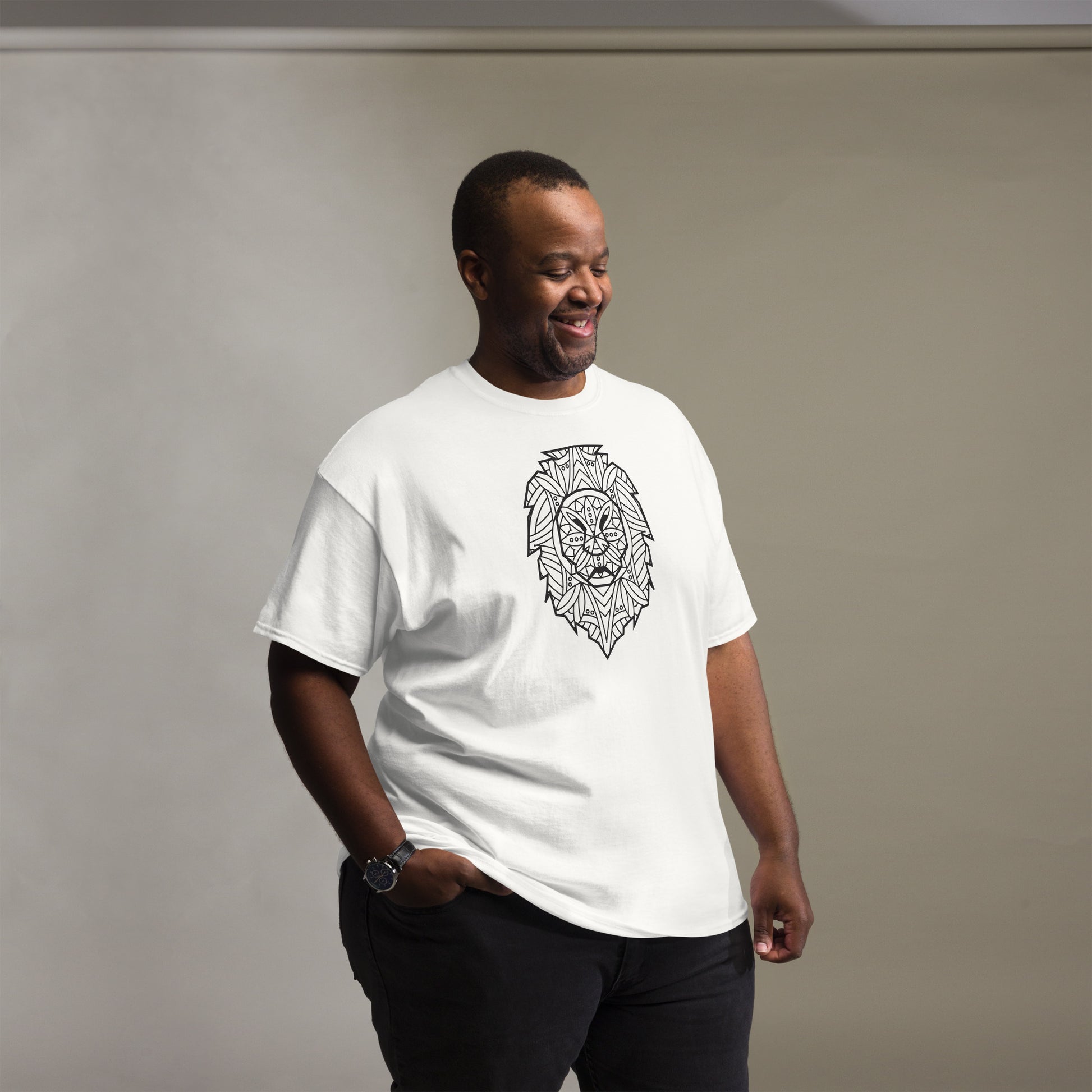 Smiling man wearing a white t-shirt with a detailed geometric lion head print on the front, paired with black pants, and a wristwatch, standing in a room with a grey background.