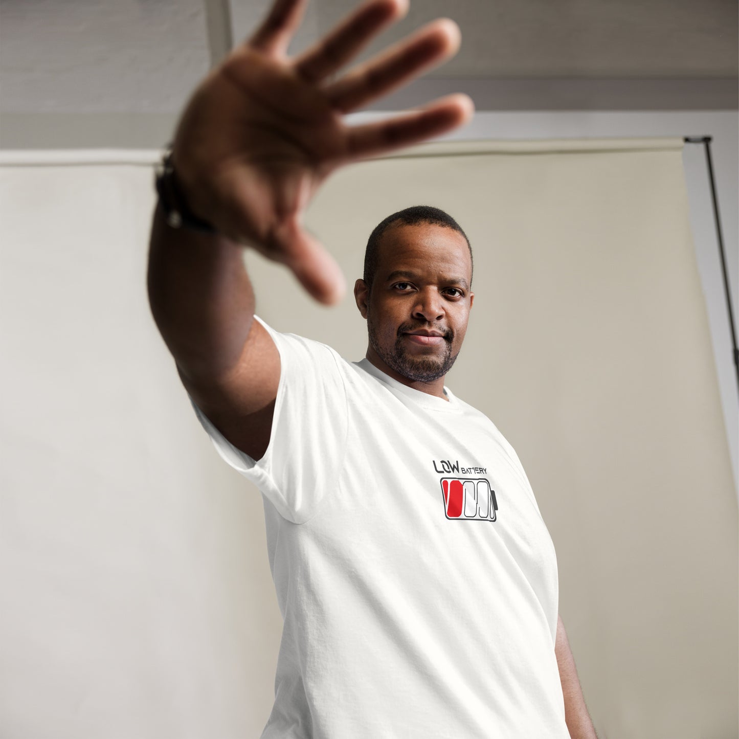 A smiling African American man poses in a white t-shirt featuring a "LOW BATTERY" design. The shirt displays a stylized battery icon with one segment filled in red, underlined by grey text with a white outline. He stands with his left hand raised towards the camera in a playful, stopping gesture. His relaxed demeanor is complemented by his casual pose and the simple, studio backdrop.